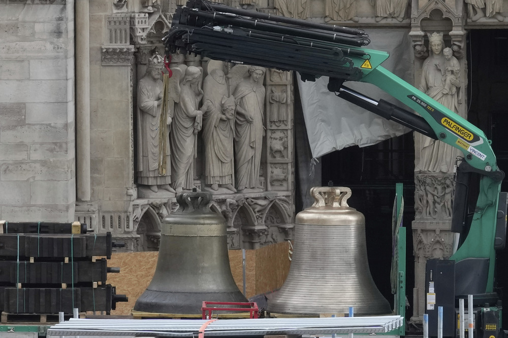 The bells of Notre Dame de Paris, Sept. 12, 2024. Large bronze bells on truck bed; in background is ornately carved portion of Cathedral exterior.