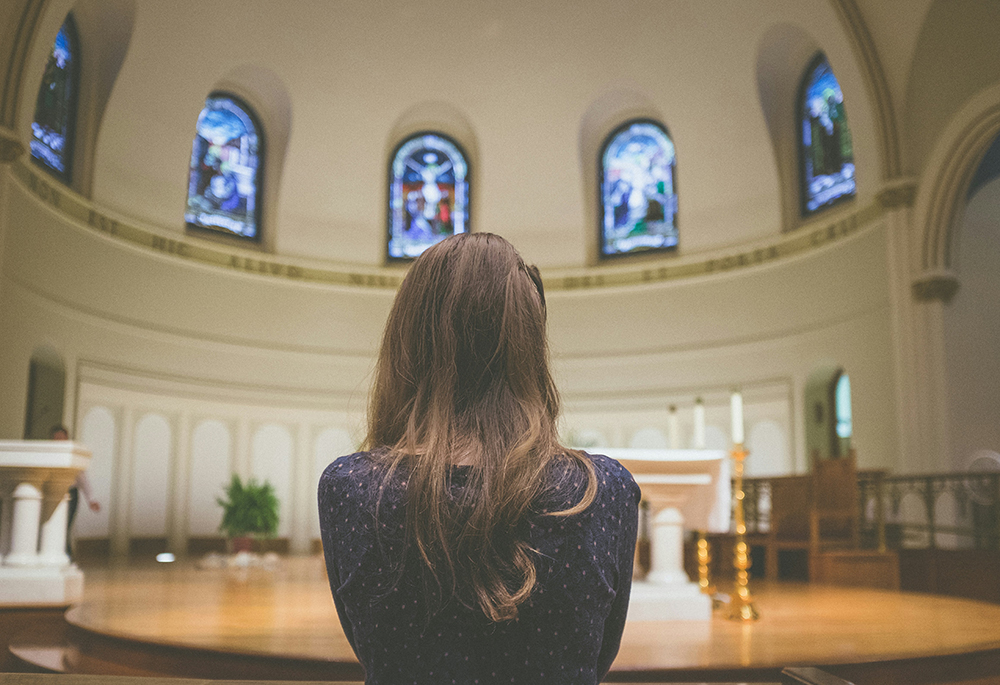 A photo shows a close-up shot of woman in the foreground, pictured inside of a church. She is facing toward an altar with her back to the viewer. (Unsplash/Kenny Eliason)