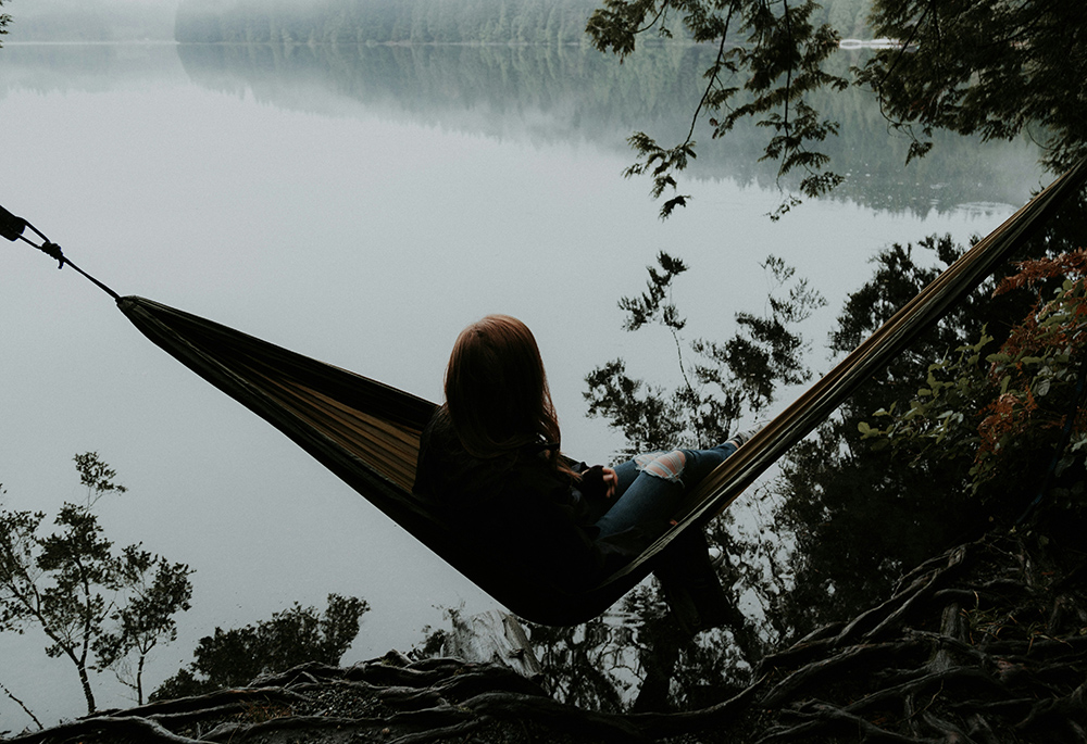 A woman sits on a hammock facing towards a lake in a forest setting (Unsplash/Priscilla Du Preez)