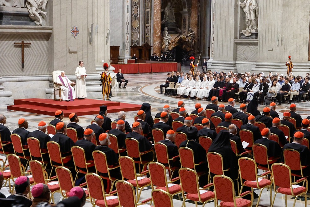 Pope seated on red dais; to his left is a group of young people, and to his right facing straight ahead are rows of prelates.