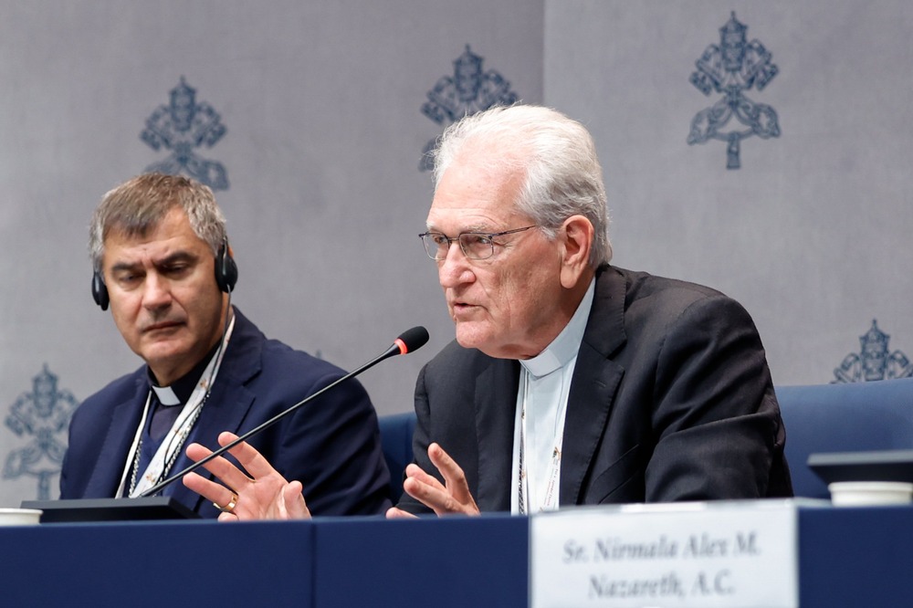 Steiner shown sitting and blue-clothed press conference table speaking into microphone; behind is a background emblazoned with the Vatican's coat-of-arms.