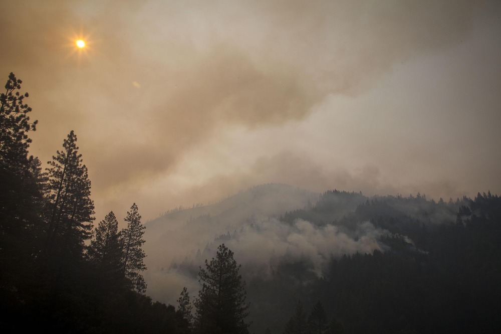 Hazy sky punctuated by sun, framed by evergreens and looming mountains.