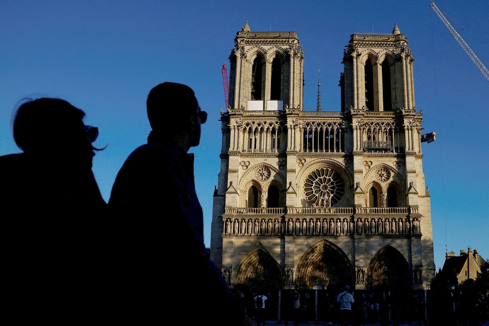 Notre Dame de Paris, reconstruction, Oct. 23, 2024. The Cathedral looms against blue sky; on right side is the arm of a crane; in left third of frame are two silhouetted figures of bystanders.