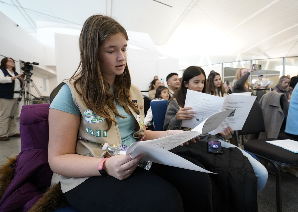 Girl wearing scout vest and holding program fills frame. Behind are other troopers singing. 