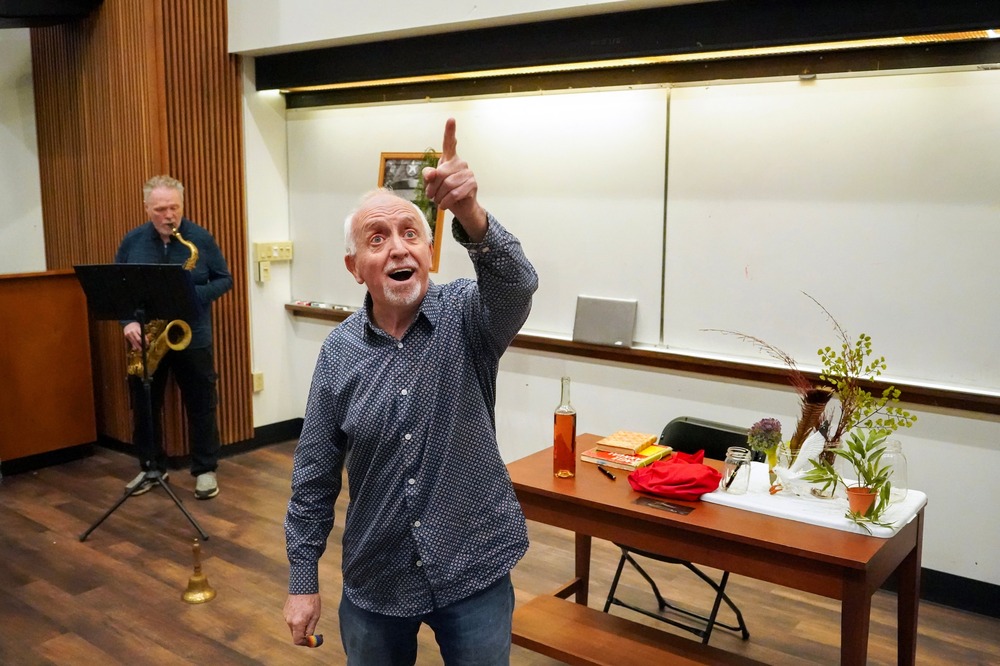 'Groomed' theater performance, Siena College, Loudonville, New York, Oct. 20, 2024. Sandford looks up and out pointing, behind him is classroom whiteboard and Benoit with saxophone.