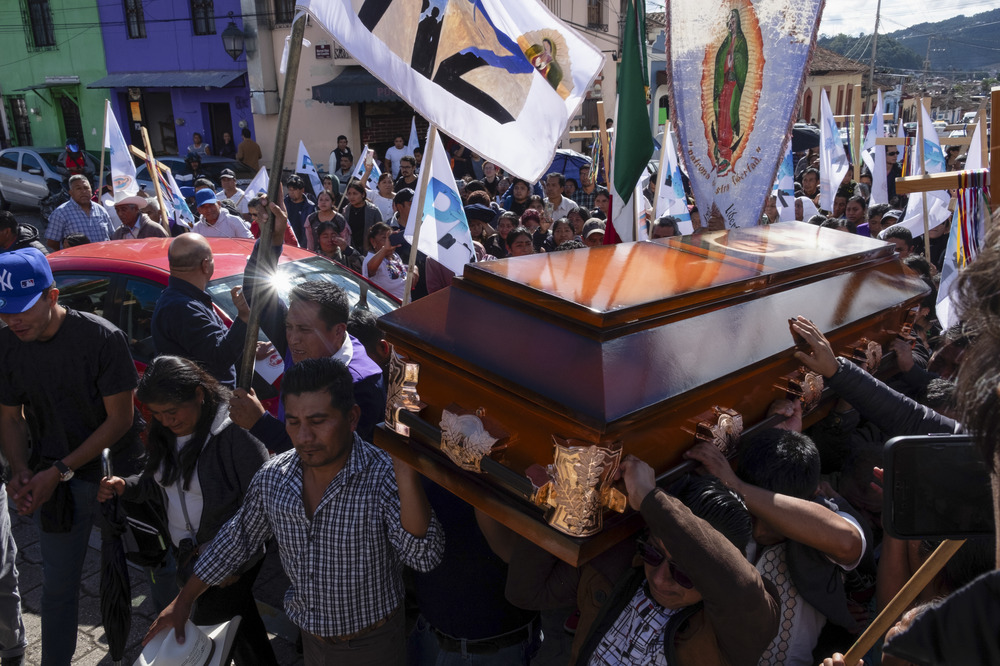 Pall-bearers carry casket, surrounded by large group carrying banners and votives.