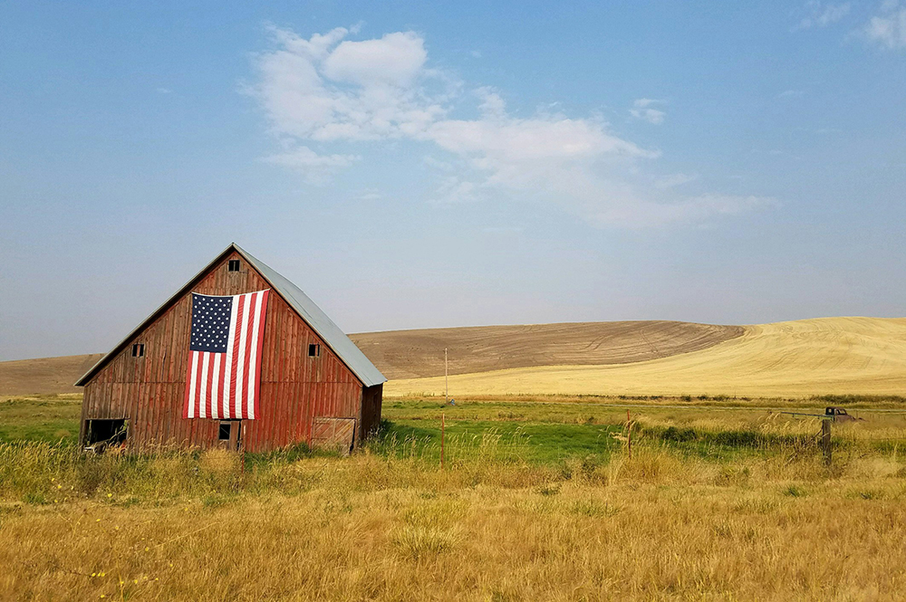 A red barn sits in the middle of a field with an American flag hanging on its side (Unsplash/specphotops)