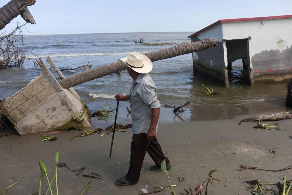 Man with walking stick foregrounded, in background are building ruins and the shoreline.