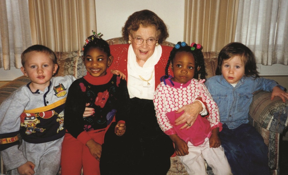 Barber sits on a couch, smiling, with two children on each side. 