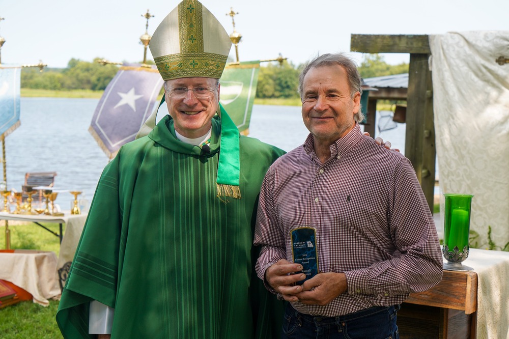 Bishop W. Shawn McKnight of Jefferson City, Mo., and Russ Kremer, Missouri farmer. The bishop, vested for ordinary time and wearing mitre, stands with his arm around Kremer who holds his award plaque.