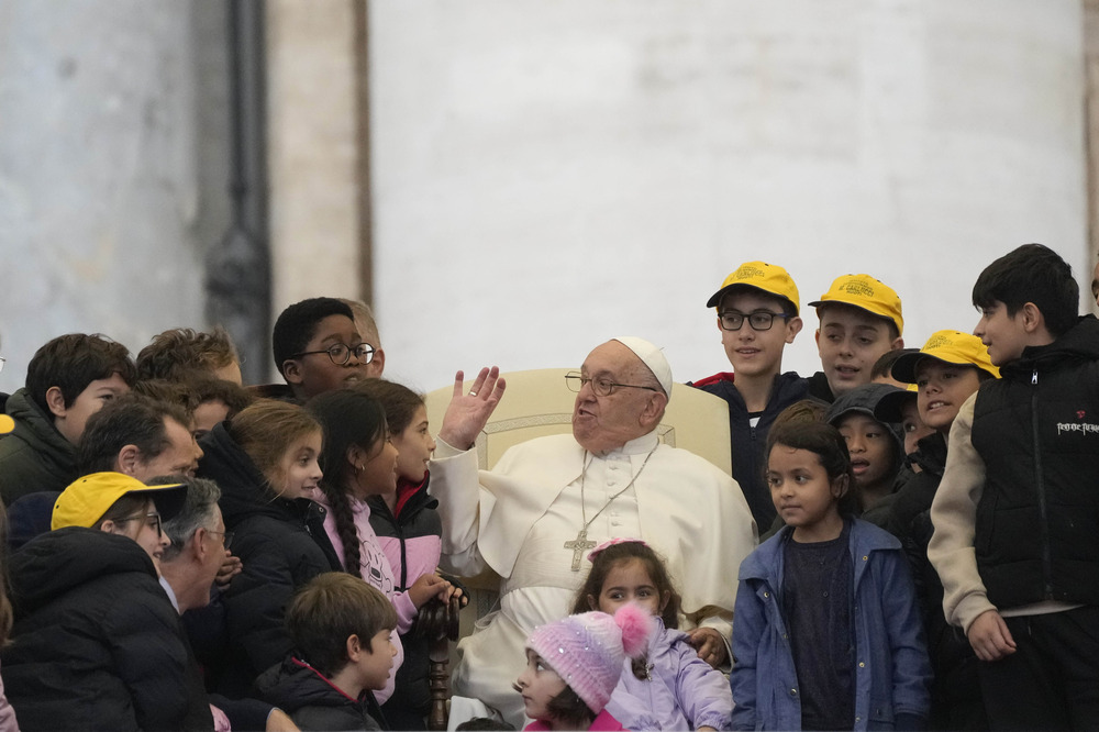 Pope sits surrounded by children; he gestures and smiles, wearing glasses.