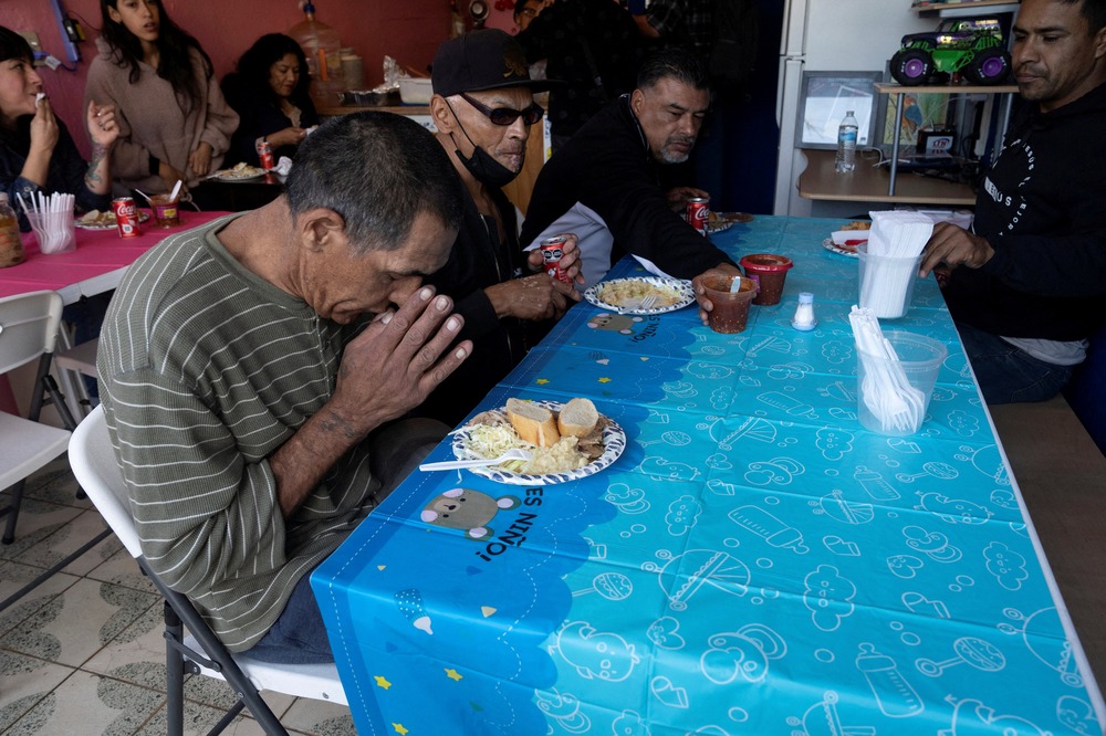 Group of people sit at long tables; foregrounded man bends his head in prayer. 