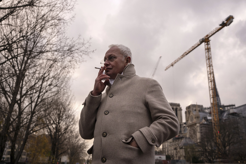 Villeneuve wears coat, smoking, in background is massive crain.