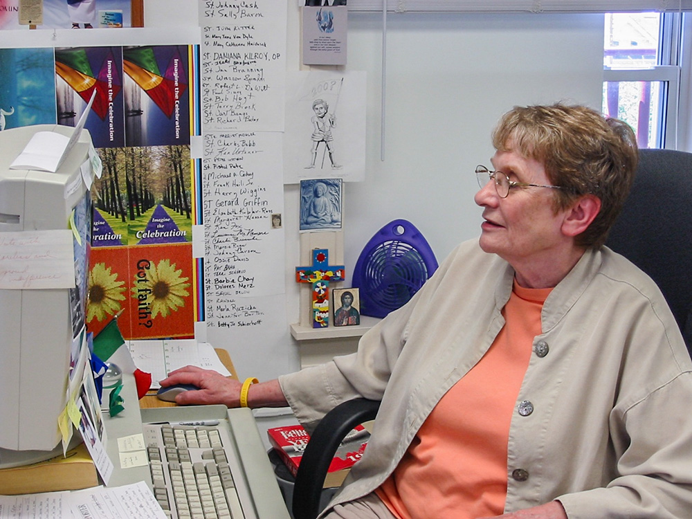 An undated photo of Patty McCarty at her NCR newsroom desk in Kansas City, Missouri. A partial list of saints is on the wall and other icons surround her. (Courtesy of Tom Fox)