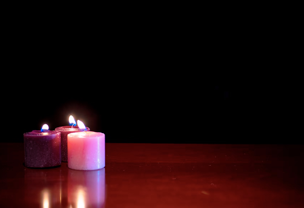 Three lit candles (two purple, one pink) are pictured on a smooth table in the dark. (Unsplash/Robert Thiemann)