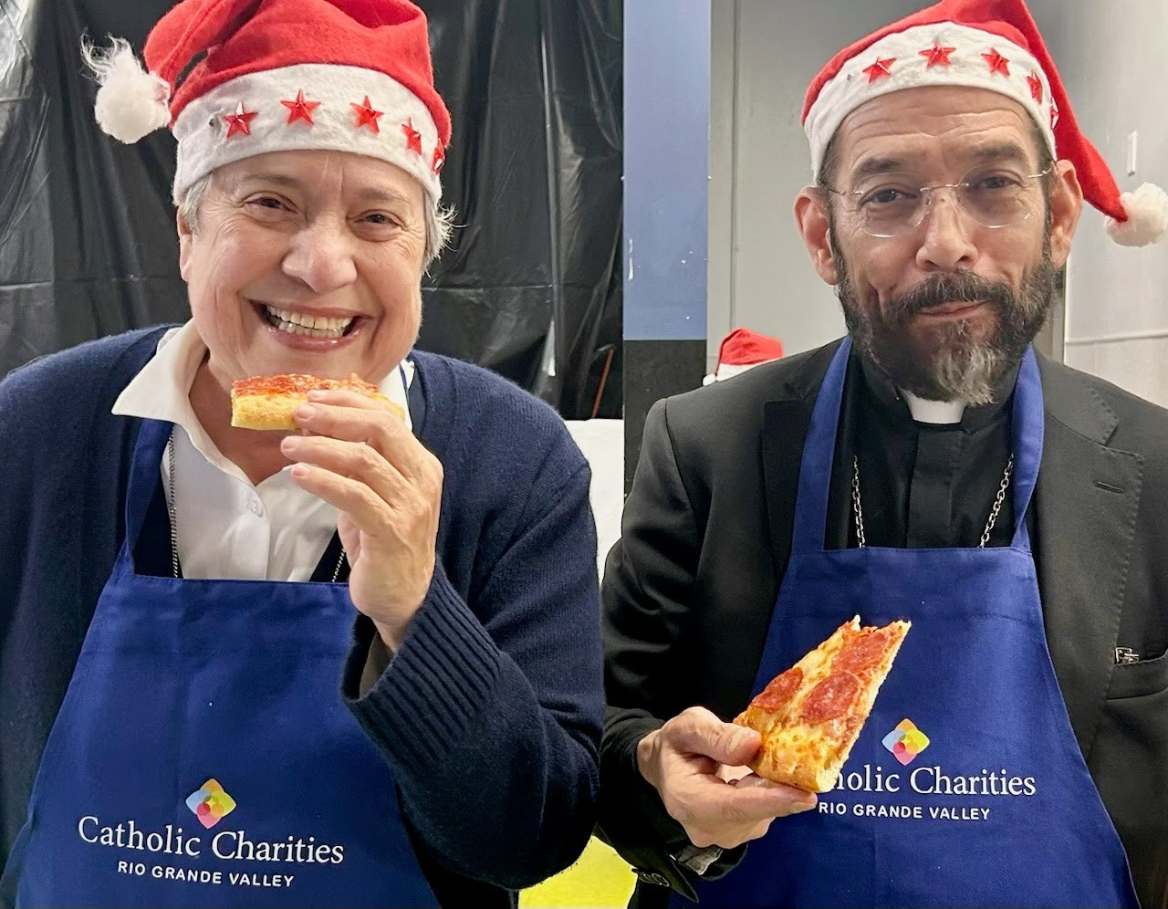 The sister and the bishop smile, wearing aprons and holding slices of pizza.