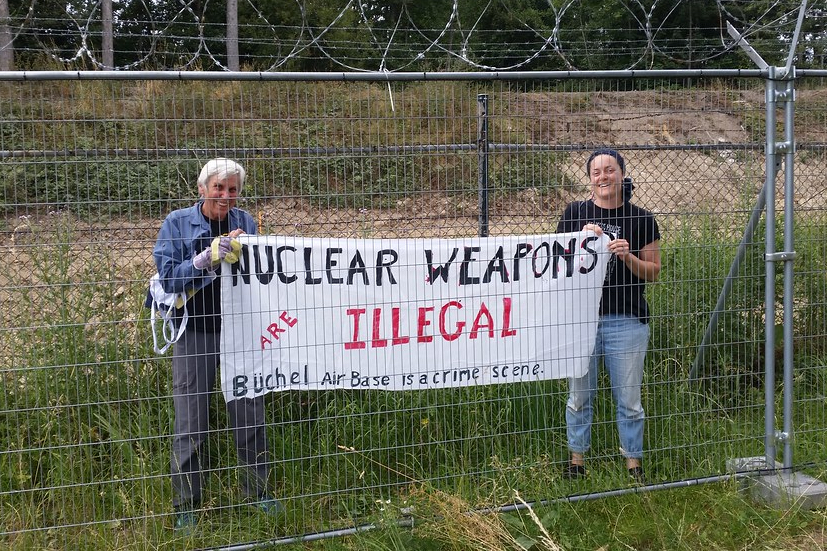 Susan Crane Büchel 1 Eighty-one-year-old activist grandma Susan Crane (left), a Catholic Worker from Redwood City, California, takes a banner with her anti-nuclear proliferation message inside Büchel Air Force Base in Büchel, Germany. The banner reads, "NUCLEAR WEAPONS ARE ILLEGAL; Büchel Air Base is a crime scene." (Courtesy of Susan Crane)