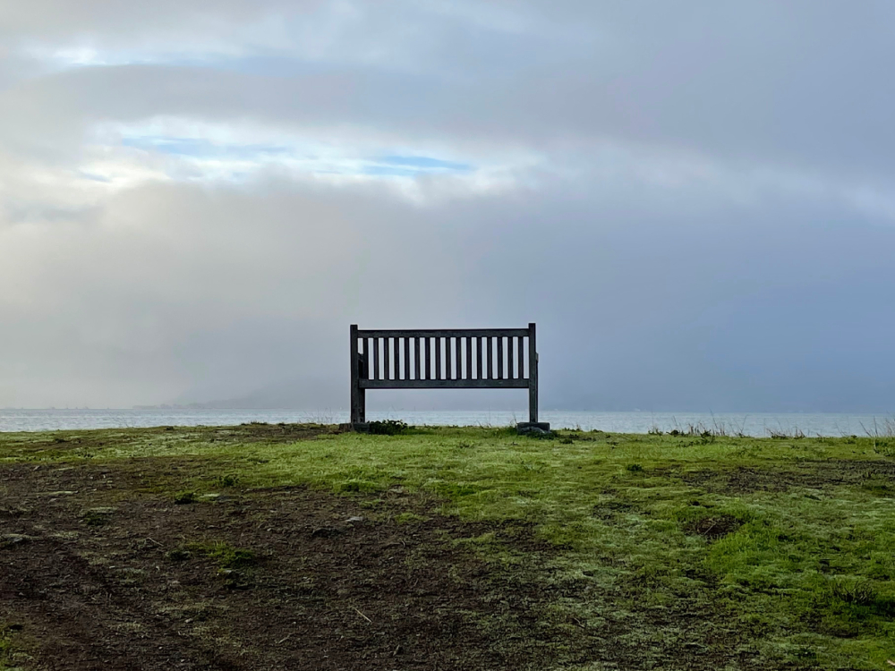 A lone bench on patchy grass overlooking a cloudy sky with patches of sunshine shining through.