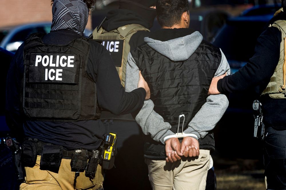 Man with Police ICE vest escorts handcuffed man.