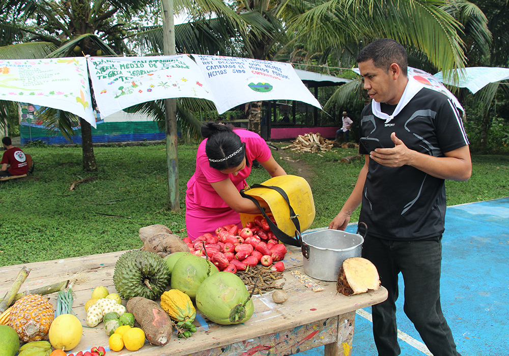 El concejal Miguel Cepeda, a la derecha, aparece en la foto con Nevaith Vargas, compartiendo los frutos de sus cosechas en una mesa común en la cancha que también sirve como lugar de encuentro comunitario para la celebración del aniversario. (Foto: Tracy L. Barnett)