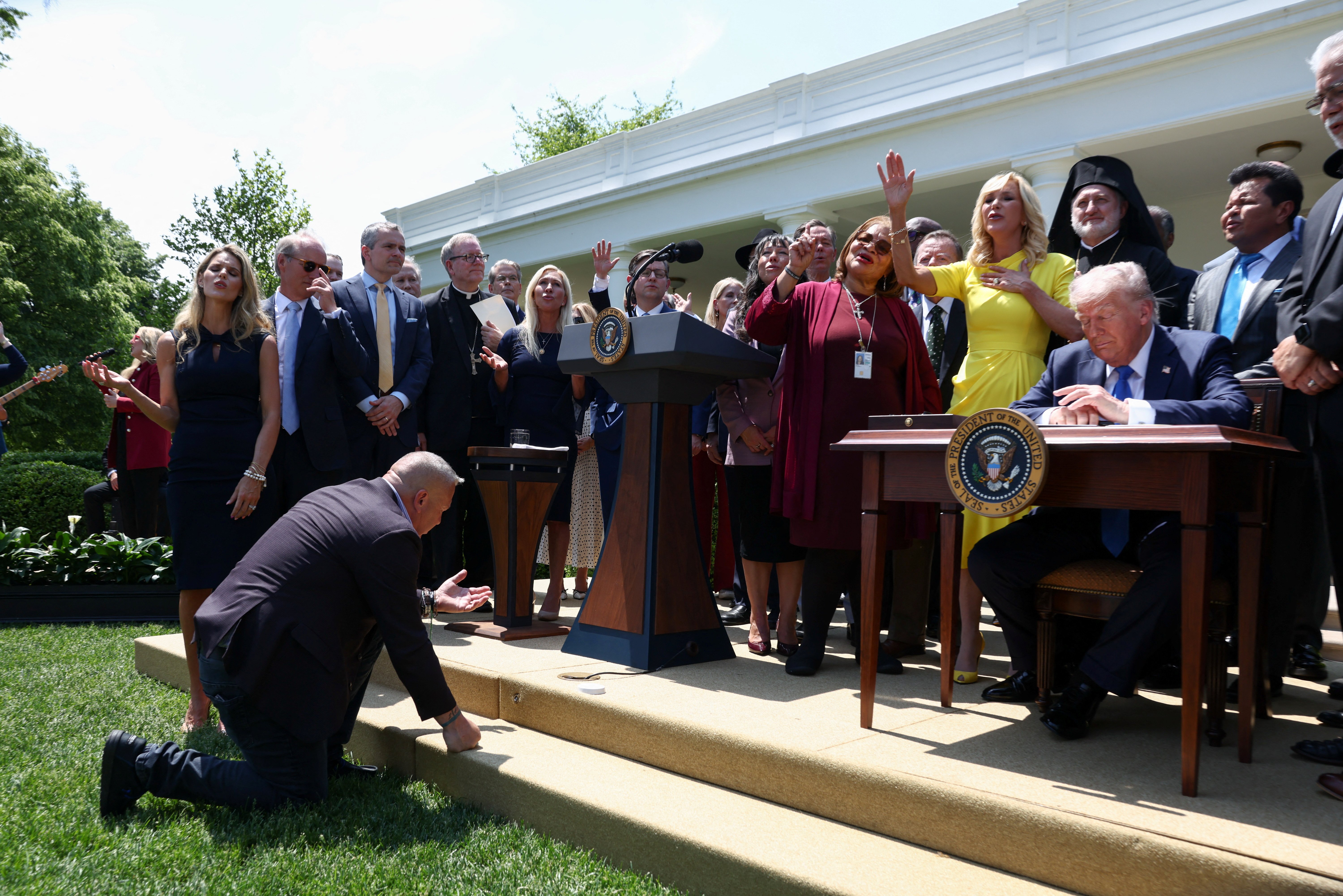An attendee kneels in front of the stage as Paula White, senior adviser to the White House Faith Office, in yellow, leads a prayer next to U.S. President Donald Trump during the National Day of Prayer in the Rose Garden at the White House in Washington May 1, 2025. Also pictured in back, fourth from left, is Bishop Robert E. Barron of Winona-Rochester, Minn. (OSV News /Evelyn Hockstein, Reuters)