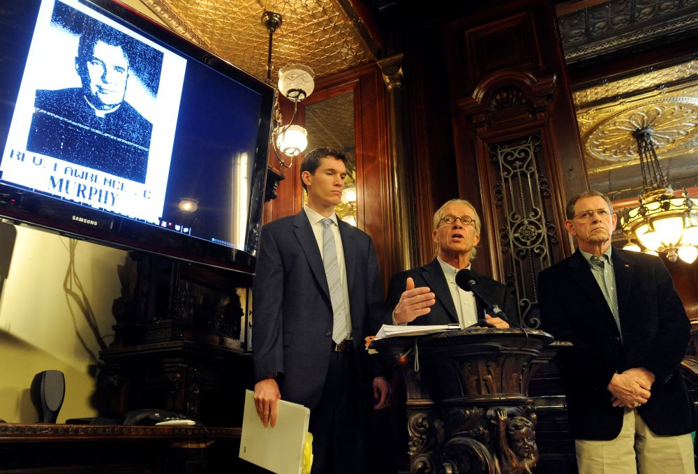 Three men at a news conference, with image of Fr. Lawrence Murphy on monitor Three men at a news conference, with image of Fr. Lawrence Murphy on monitor