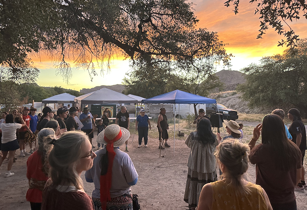 A crowd, including sisters, listening to an Apache Stronghold ally and artist rap during recent Oak Flat demonstrations. Among those who spoke at the gathering was Wendsler Nosie, an Apache elder and leader of Apache Stronghold. Nosie told the visitors of the need "to stand together" and find common ground among different groups. (Courtesy of Land Justice Features)