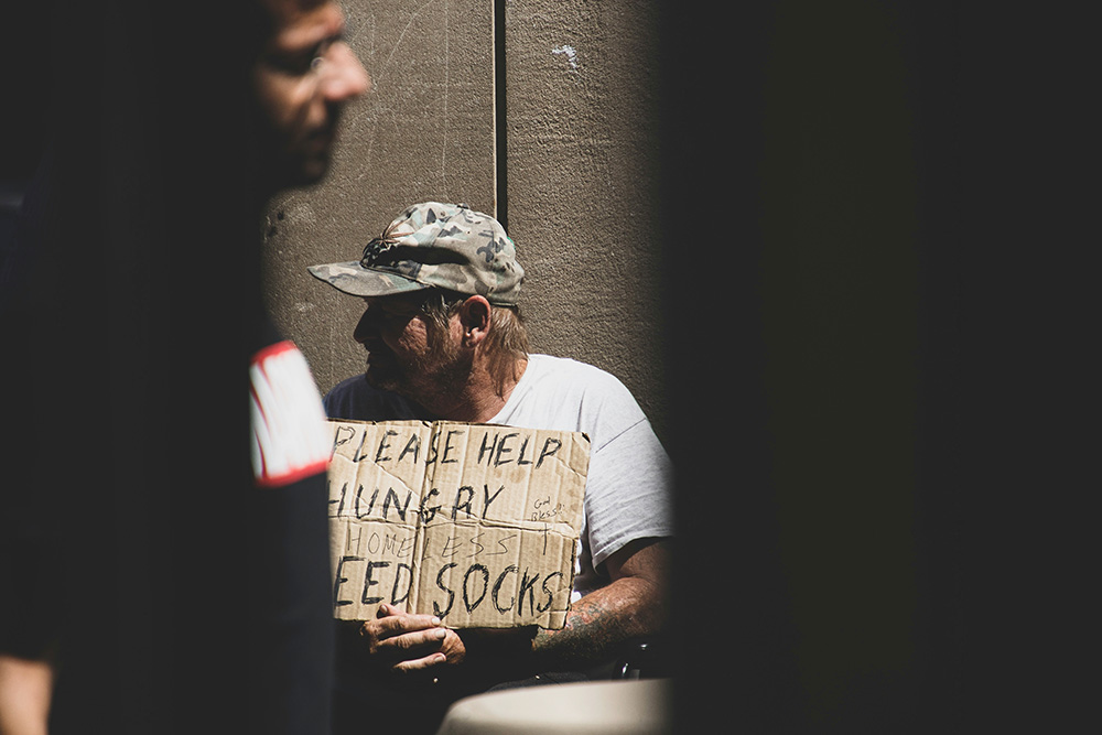 A person holds a sign saying "Please help, hungry" while another person walks by (Unsplash/Logan Weaver)