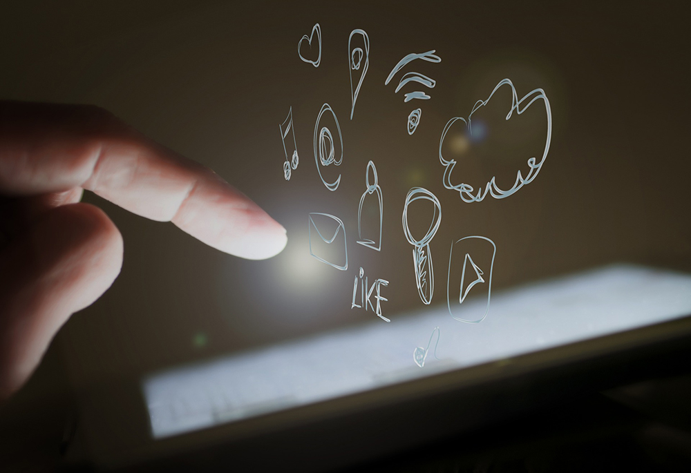 A photo illustration show a hand hovering over a smart device, with drawings of social media buttons and tags floating above the screen. (Pixabay/Edar)