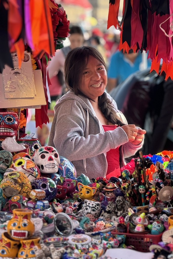 Una adolescente vende artesanías de calaveras decoradas y vasijas con imágenes de animales durante el Desfile de las Catrinas el 26 de octubre de 2025 en la Ciudad de México. (Foto: Helga Leija/GSR en español)
