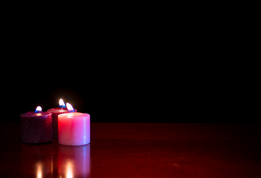 Three lit candles, two purple and one pink, burn on top of brown table in a dark room. (Unsplash/Robert Thiemann)