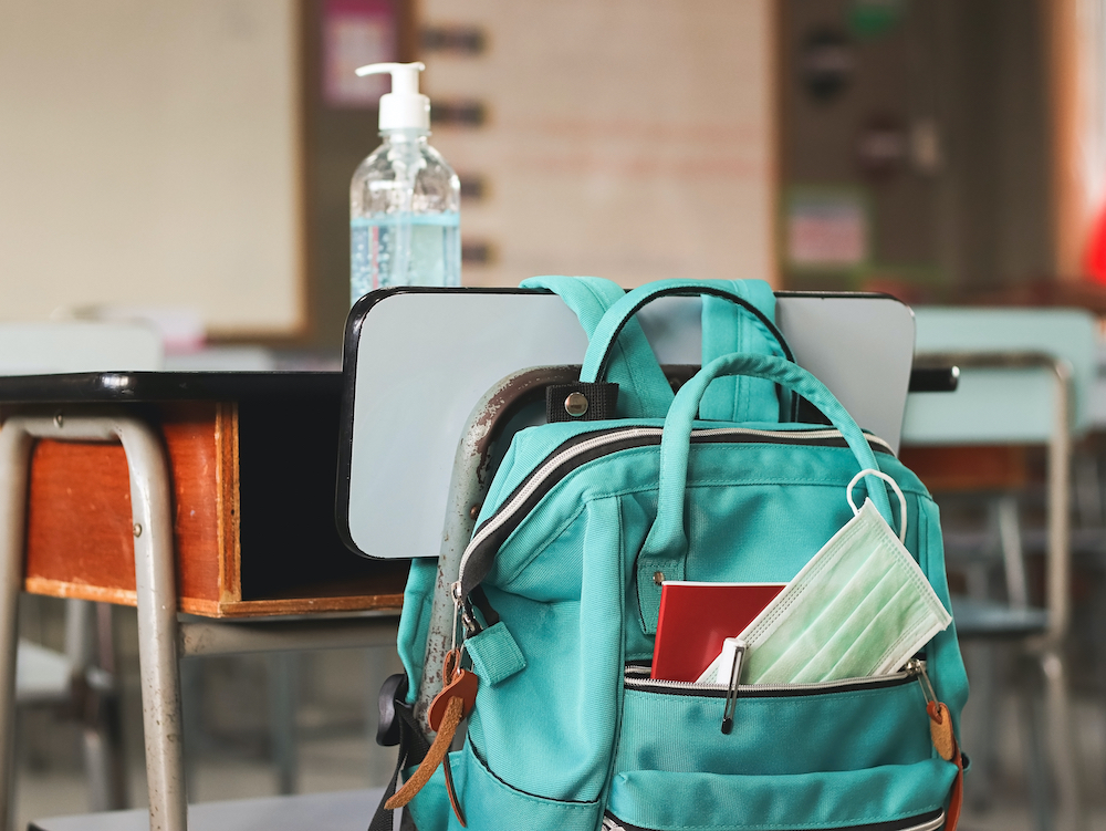 A disposable face mask is seen in a backpack hanging on a school desk holding a bottle of hand sanitizer. (Dreamstime/Phuttharak Chindarot)