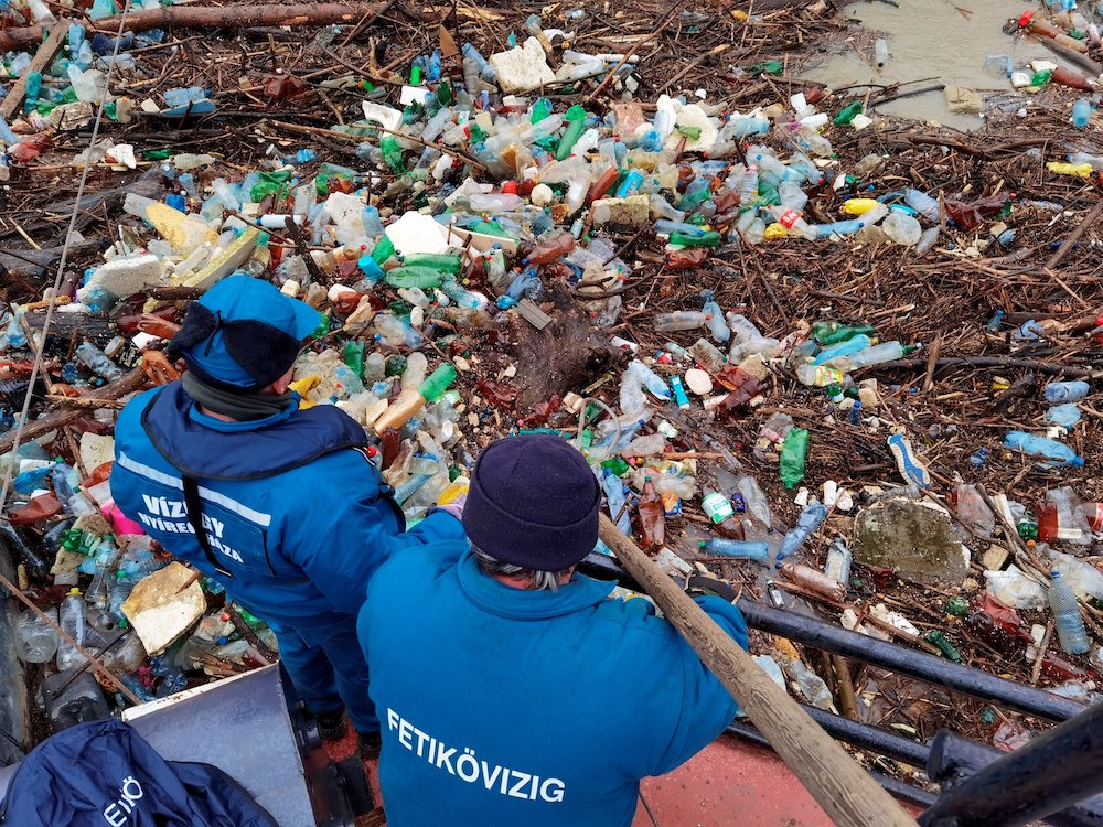 Backs of two people in a boat facing floating plastic bottles and sticks in a river