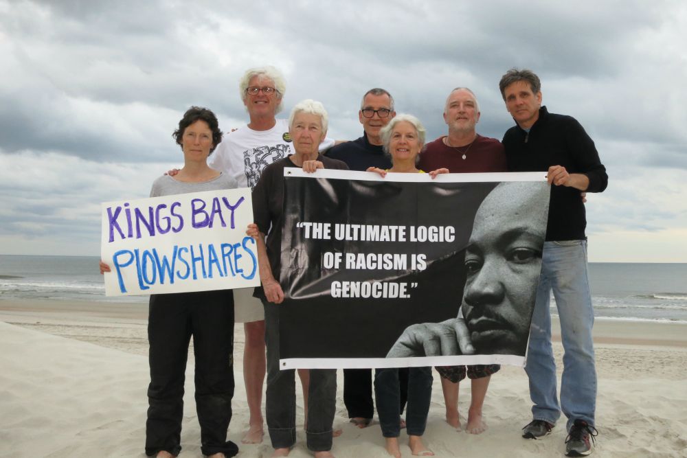 Seven people on a beach holding signs; one says "Kings Bay Plowshares," the other features a portrait of Martin Luther King, Jr. 