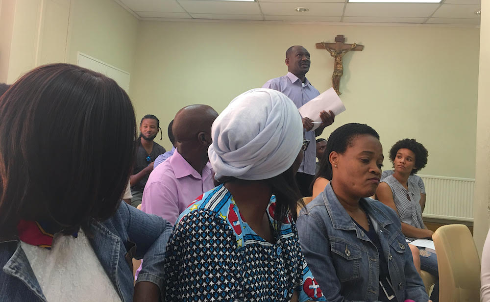 People in a room with a crucifix on the wall, one man standing up to ask a question