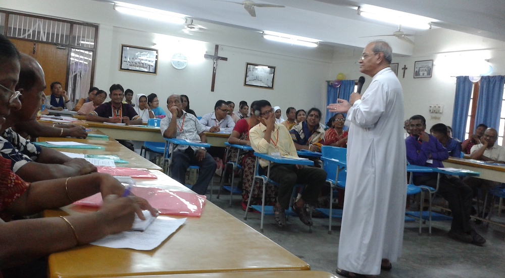 A cardinal, priest in white with a microphone addresses people seated at tables arranged around him