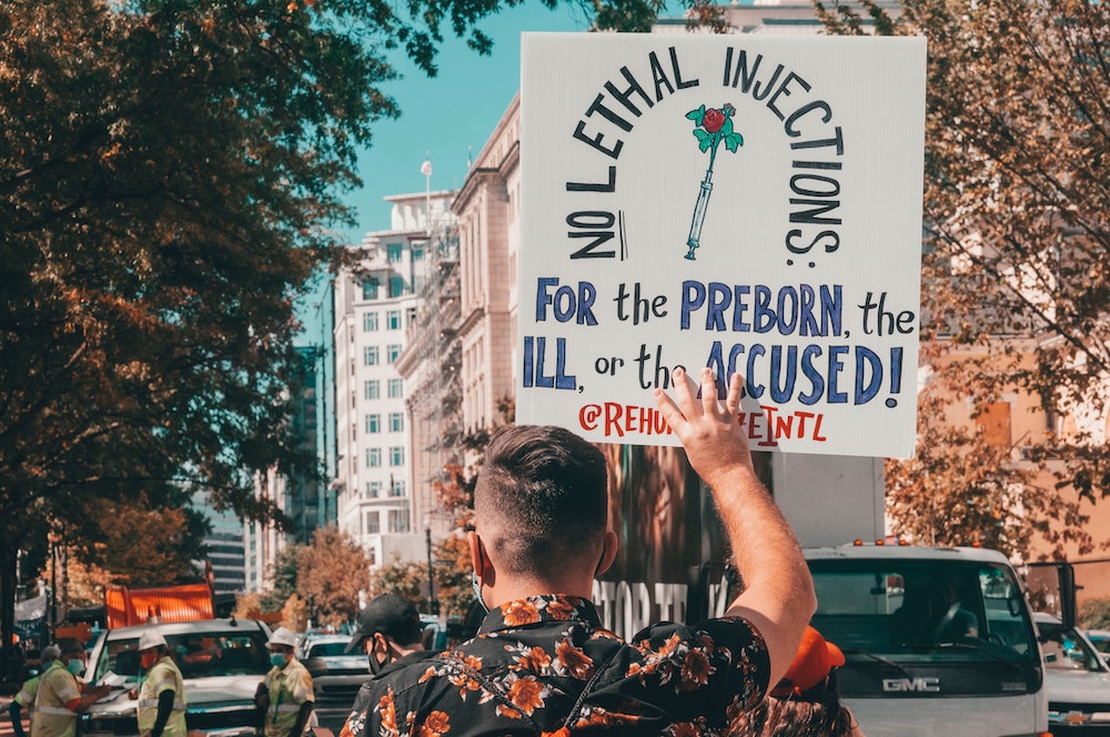 View of back of person holding sign that reads "No lethal injections for the preborn, the ill or the accused" (Unsplash Maria Oswalt)