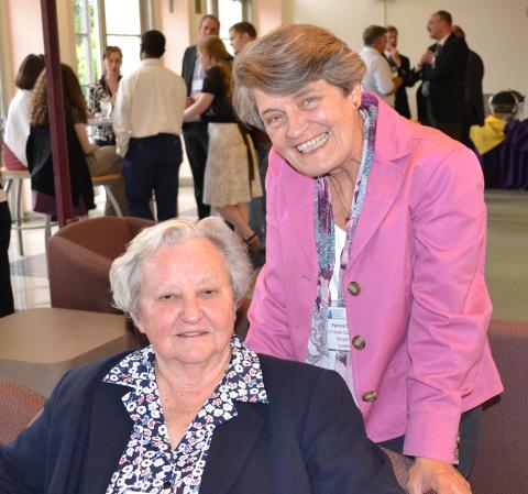 Two women smile for the camera. One is seated and the other is standing