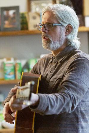 Maher stands strumming guitar. 