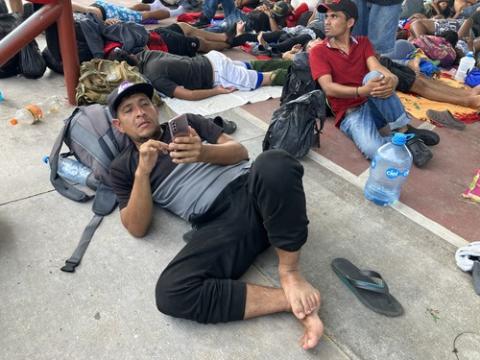 Luis lies on the ground, using his backpack as a pillow, looking at his phone. Around him filling the frame are many other migrants.