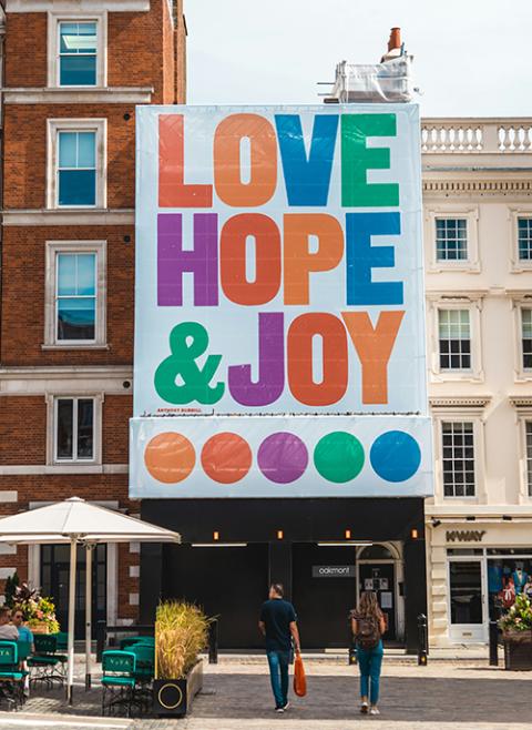 A large banner hangs from a rooftop, displaying in multicolored capital letters "LOVE HOPE & JOY." (Unsplash/Nicolas J Leclercq)