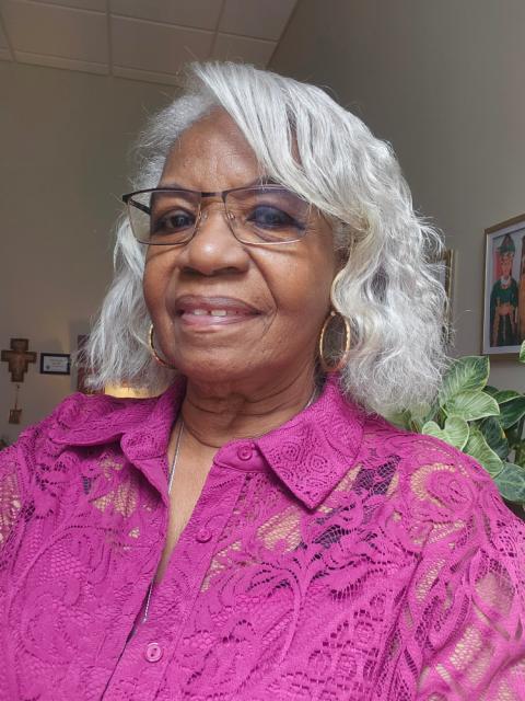 Sr. Jannette Pruitt has light grey curly, wavy hair that reaches her neck and is wearing a magenta button-up shirt and thin-rimmed square glasses.
