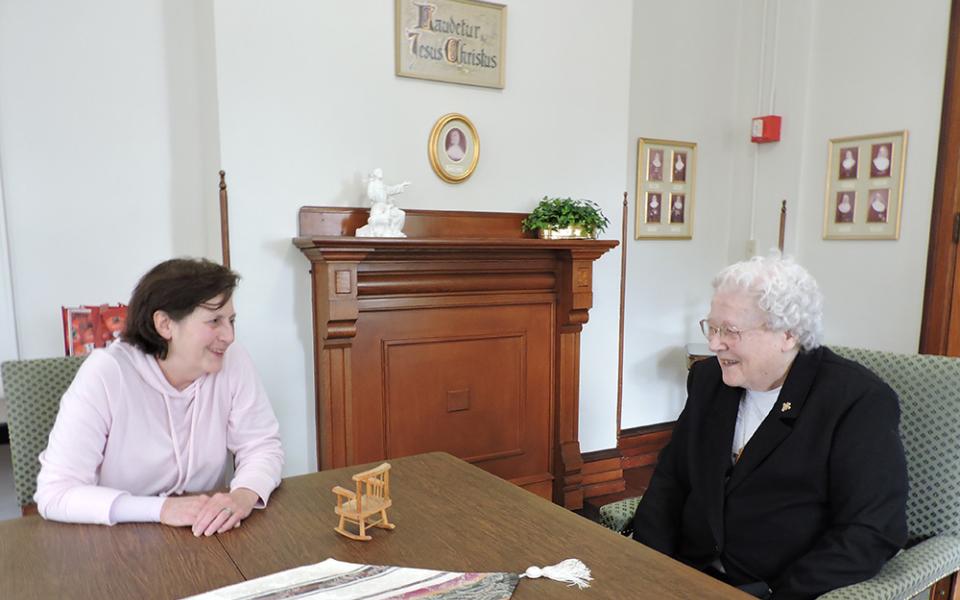 Dos religiosas conversan sentadas y apoyadas en una mesa marrón en el salón blanco de visitas del Convento de Nuestra Señora de los Ángeles, en Pensilvania.