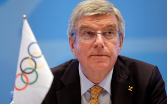 Thomas Bach sits behind a small flag with the Olympic rings