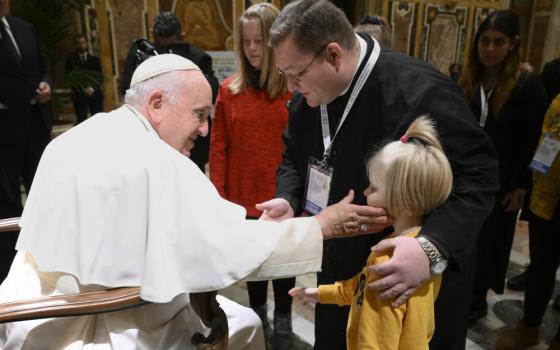 Pope Francis sits in a chair and touches a child's cheek as the child's father stands beside them