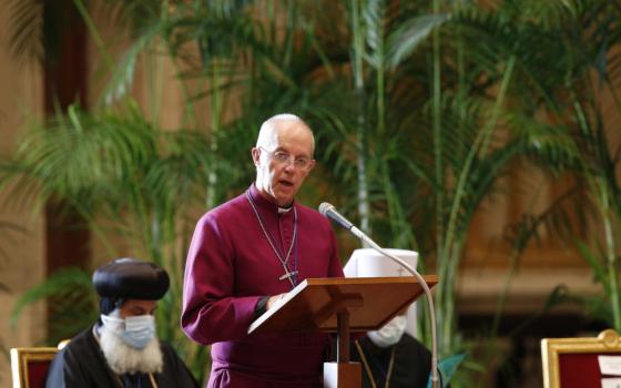 Justin Welby, an older white man wearing glasses, a cross, and a magenta clergy shirt, speaks into a microphone with large plants behind him