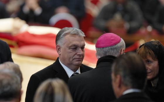 A gray-haired man in a suit talks to a bishop seen from the back with Pope Benedict XVI's body in the background