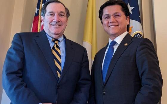 An older white man in a suit stands next to a Brown-skinned man in a suit