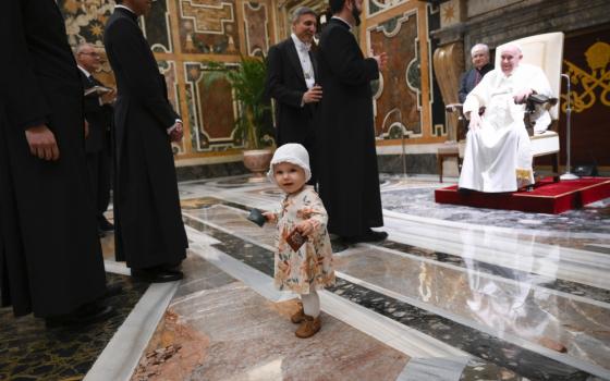 A toddler with a white hat stands in front of men in cassocks who wait to greet Pope Francis