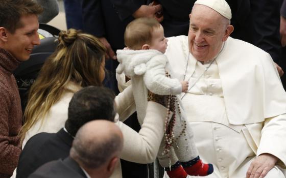 Pope Francis smiles at a baby who is held up towards him by a woman who also clutches many rosaries to the baby's side